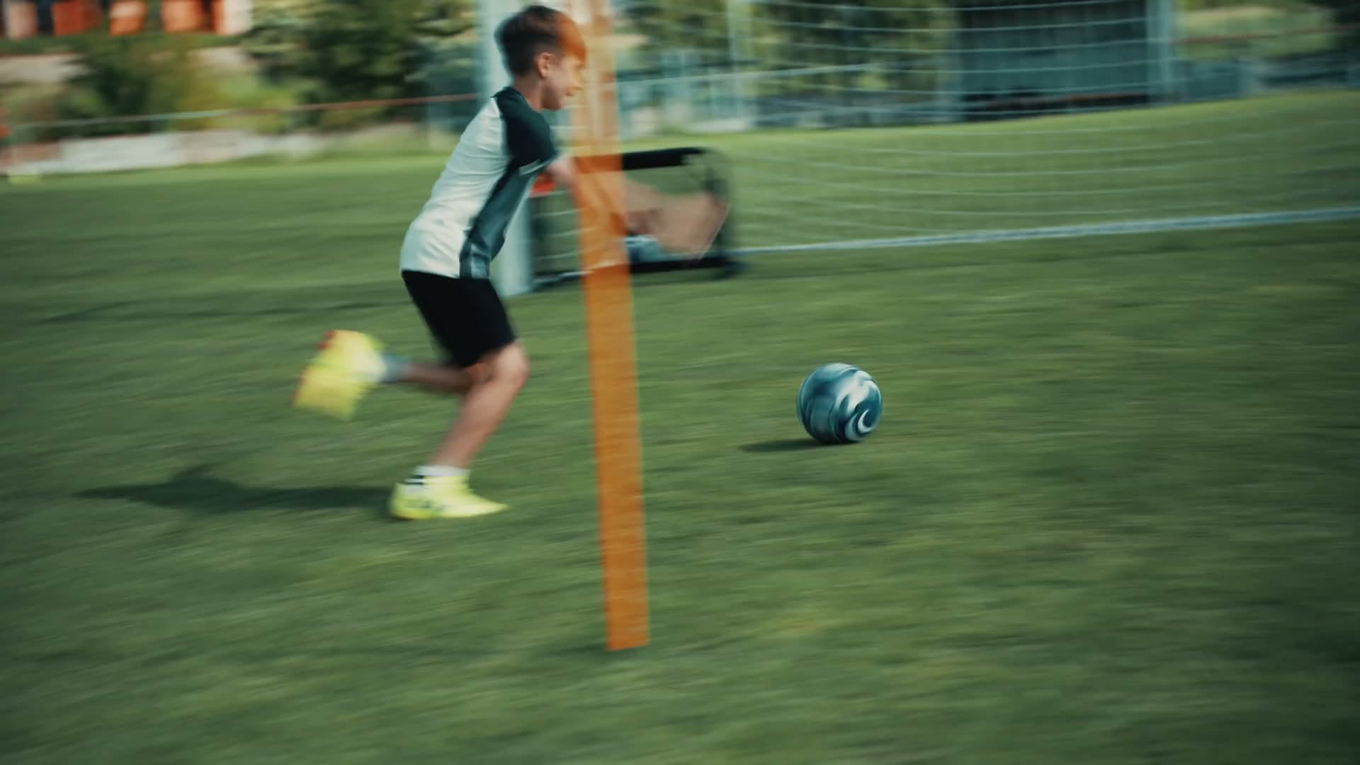 Mladý hráč zrýchľuje s loptou pri tréningovej tyči na futbalovom ihrisku.